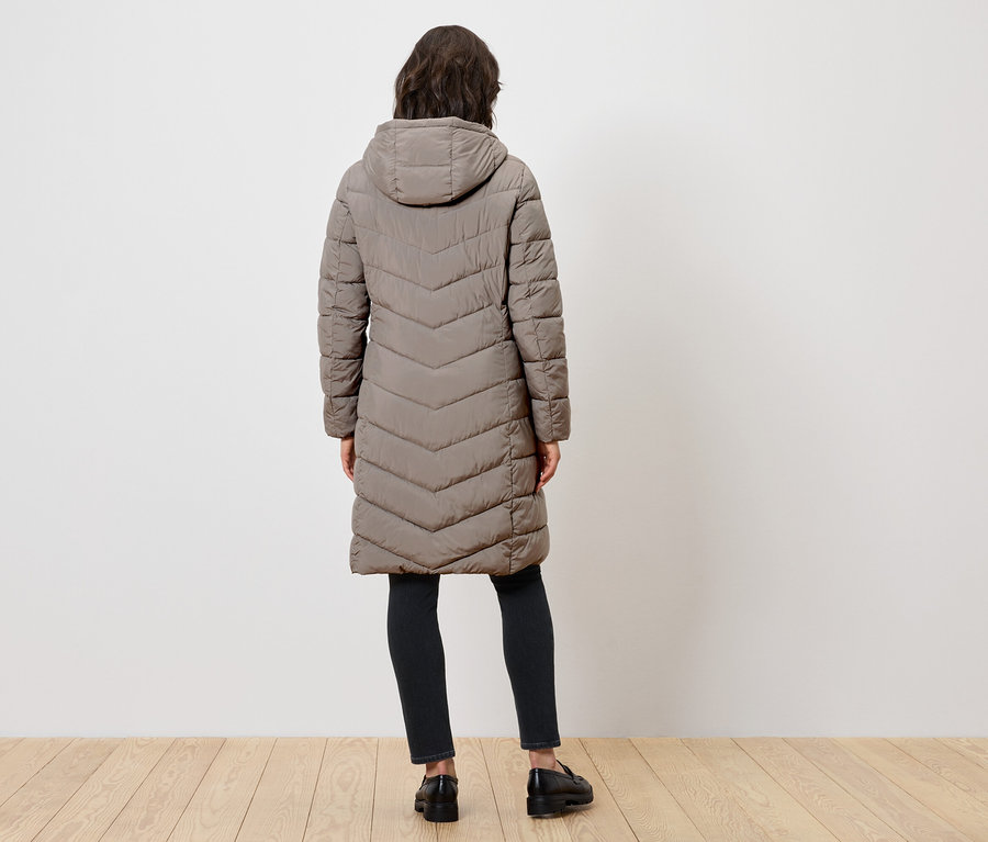 Žena chrbtom k fotoaparátu má na sebe dlhý prešívaný kabát s kapucňou, čierne nohavice a čierne topánky.