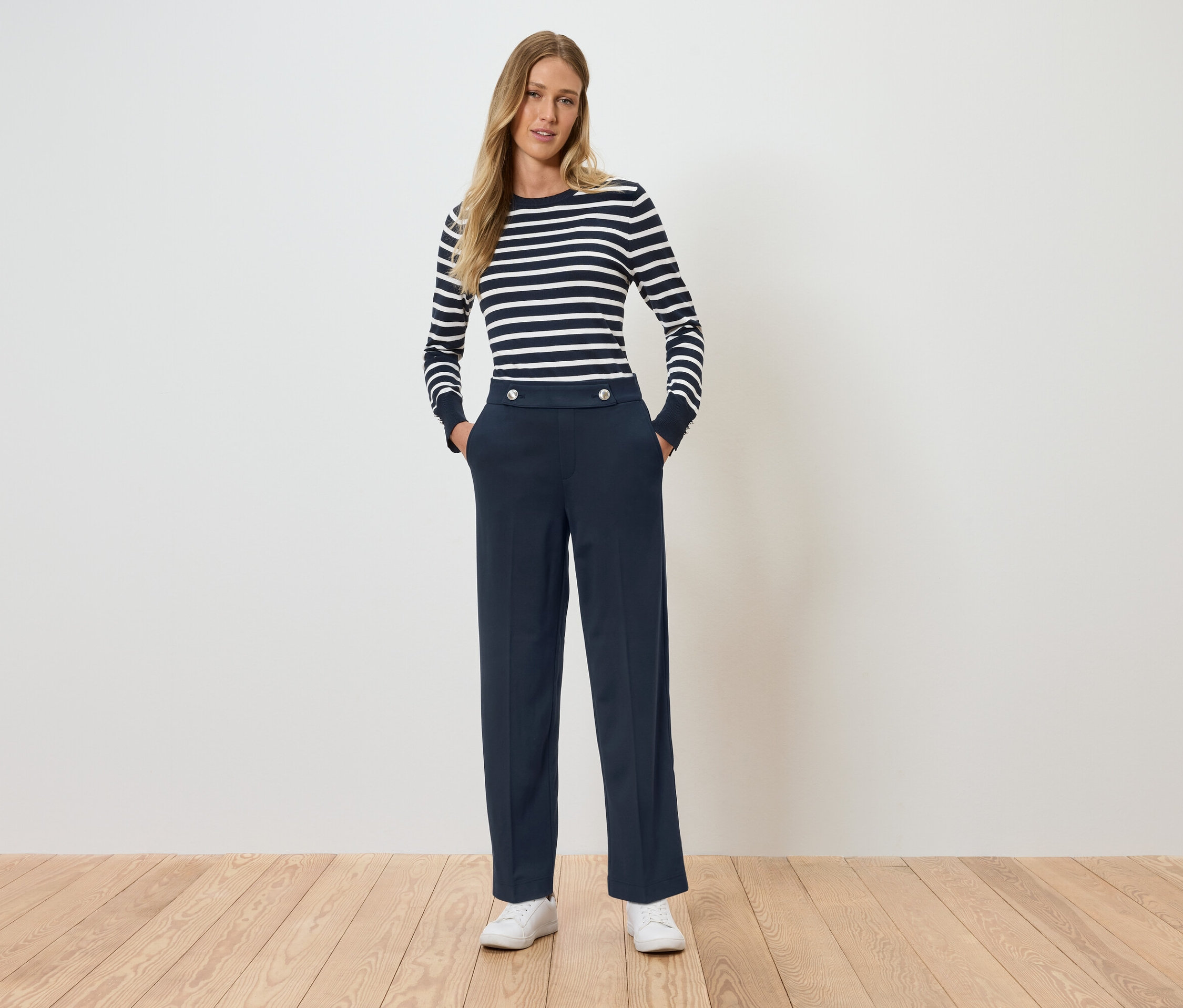 Žena stojí a má ruky vo vreckách. Má na sebe pruhované tričko s dlhým rukávom a tmavomodré nohavice.