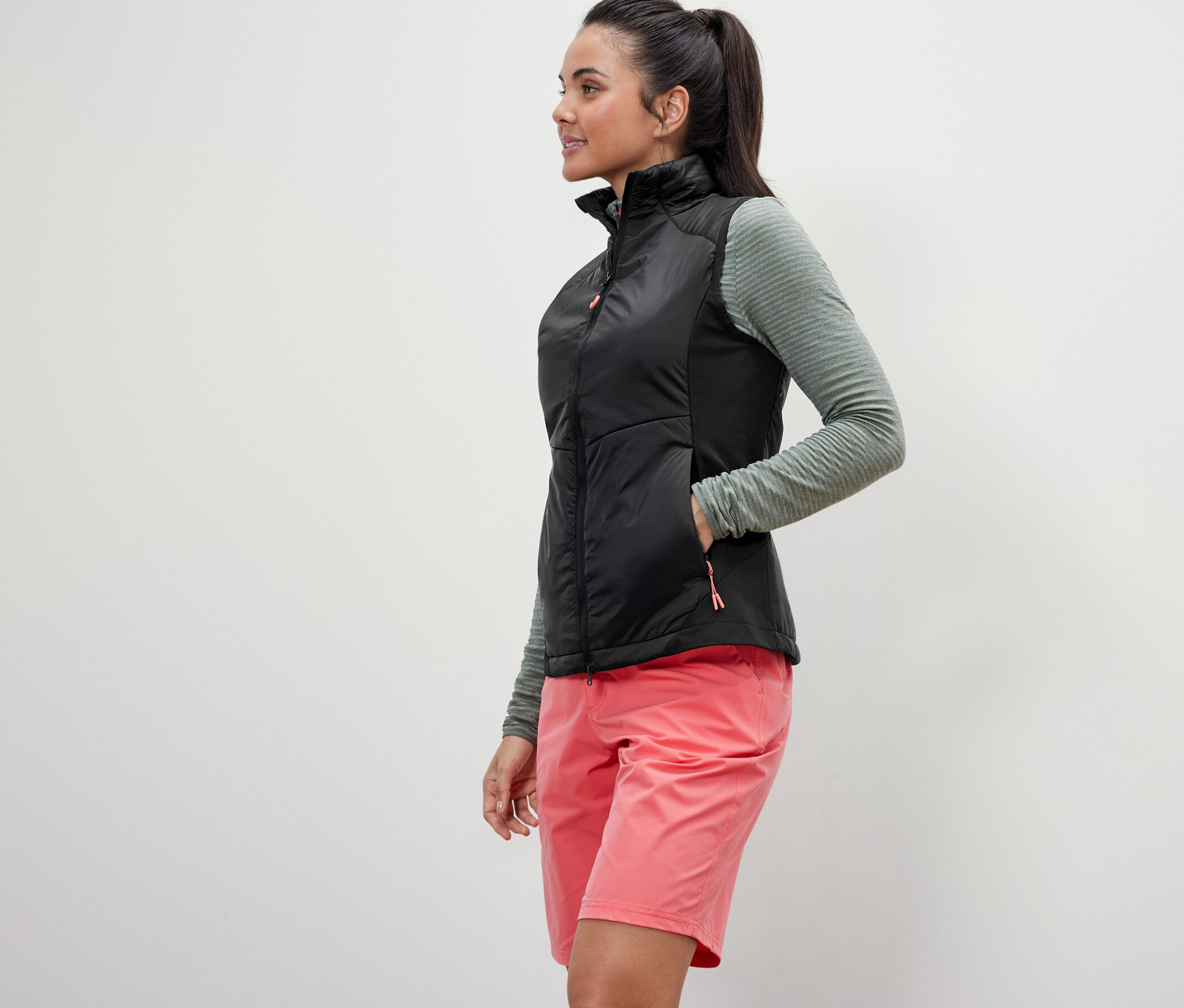 Žena pózuje v hybridnej veste »pack light«, olivovom tope a ružových šortkách.