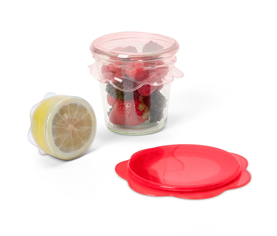 Pohár s ovocím zakrytý 3 silikónové veká na udržiavanie čerstvosti, vedľa neho polovica citróna zakrytá rovnakým vekom a červené veko.