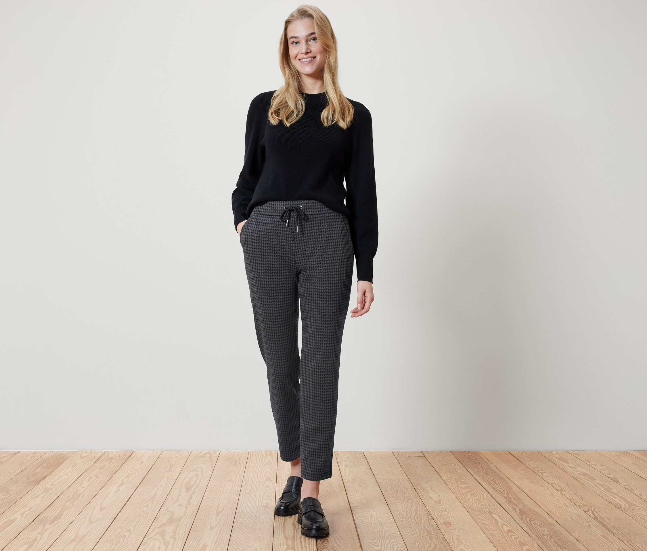 Žena stojí a má ruky vo vreckách nohavíc s kockovaným vzorom. Má na sebe čierny sveter a čierne topánky.
