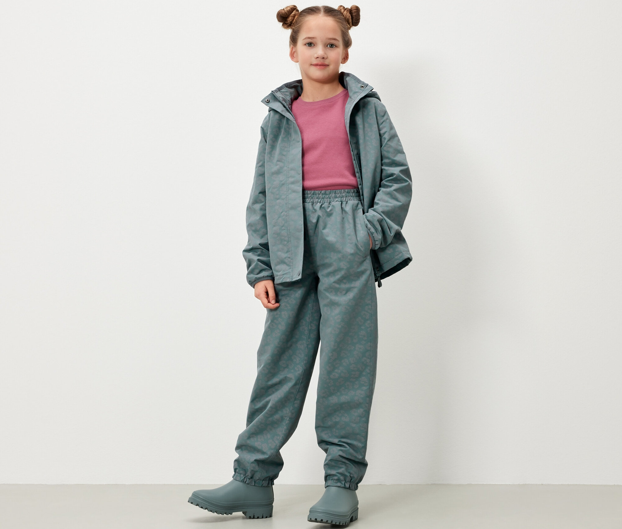 Dievča stojí a pózuje v detských nohaviciach do dažďa s reflexnými prvkami, bunde a gumákoch v sivozelenej farbe.