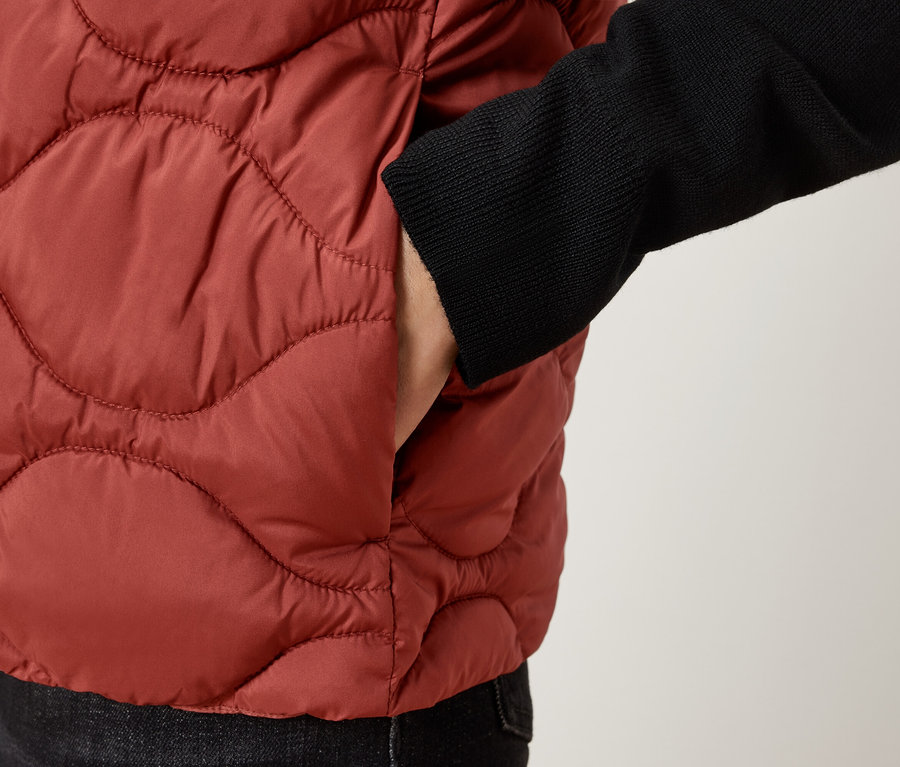 Detailný záber na osobu v hnedej prešívanej veste s rukou vo vrecku.