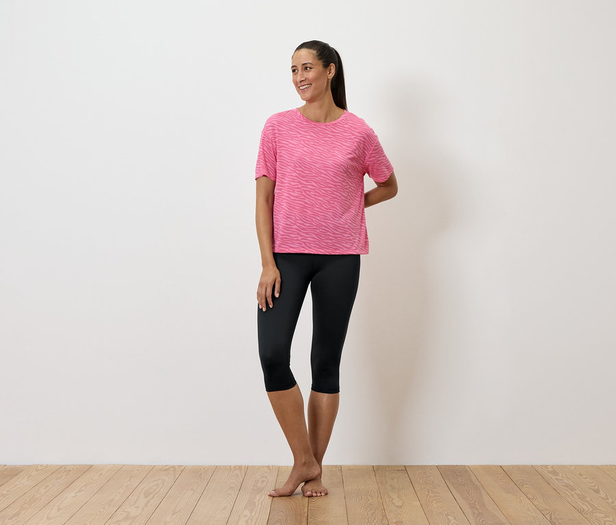 Žena stojí a má na sebe ružové tričko a čierne legíny.