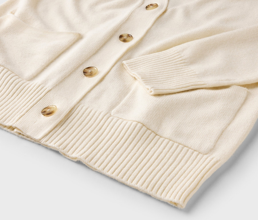 Detail krémového svetra so zapínaním na gombíky a vreckami.