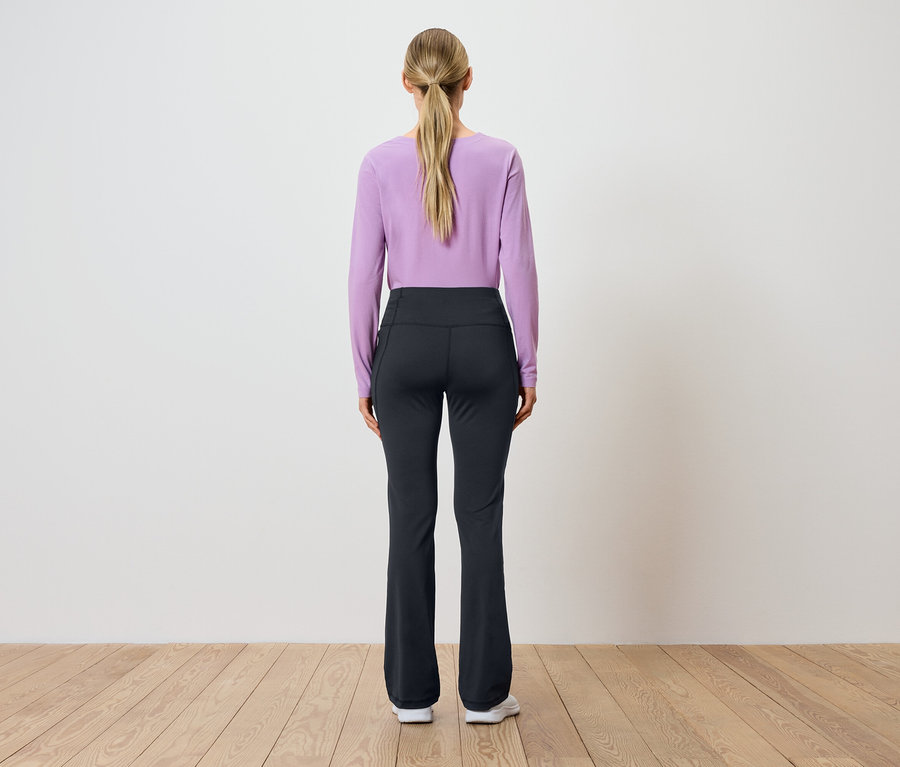 Pohľad zozadu na ženu s blond vlasmi stiahnutými do copu, oblečenú v fialovom tope s dlhým rukávom a čiernych nohaviciach.