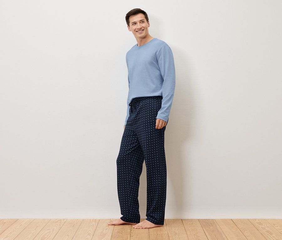 Muž stojí bosý pred bielou stenou a má na sebe svetlomodrý pulóver a tmavomodré pyžamové nohavice s bodkovaným vzorom.
