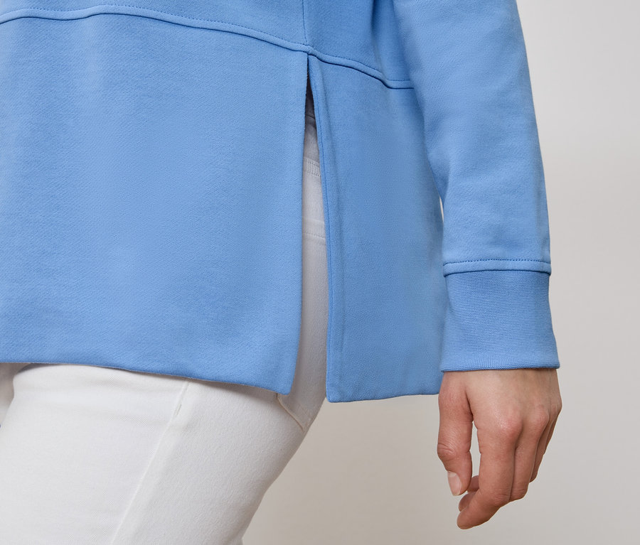 Osoba má na sebe svetlomodrú mikinu s bočným rozparkom a biele nohavice.