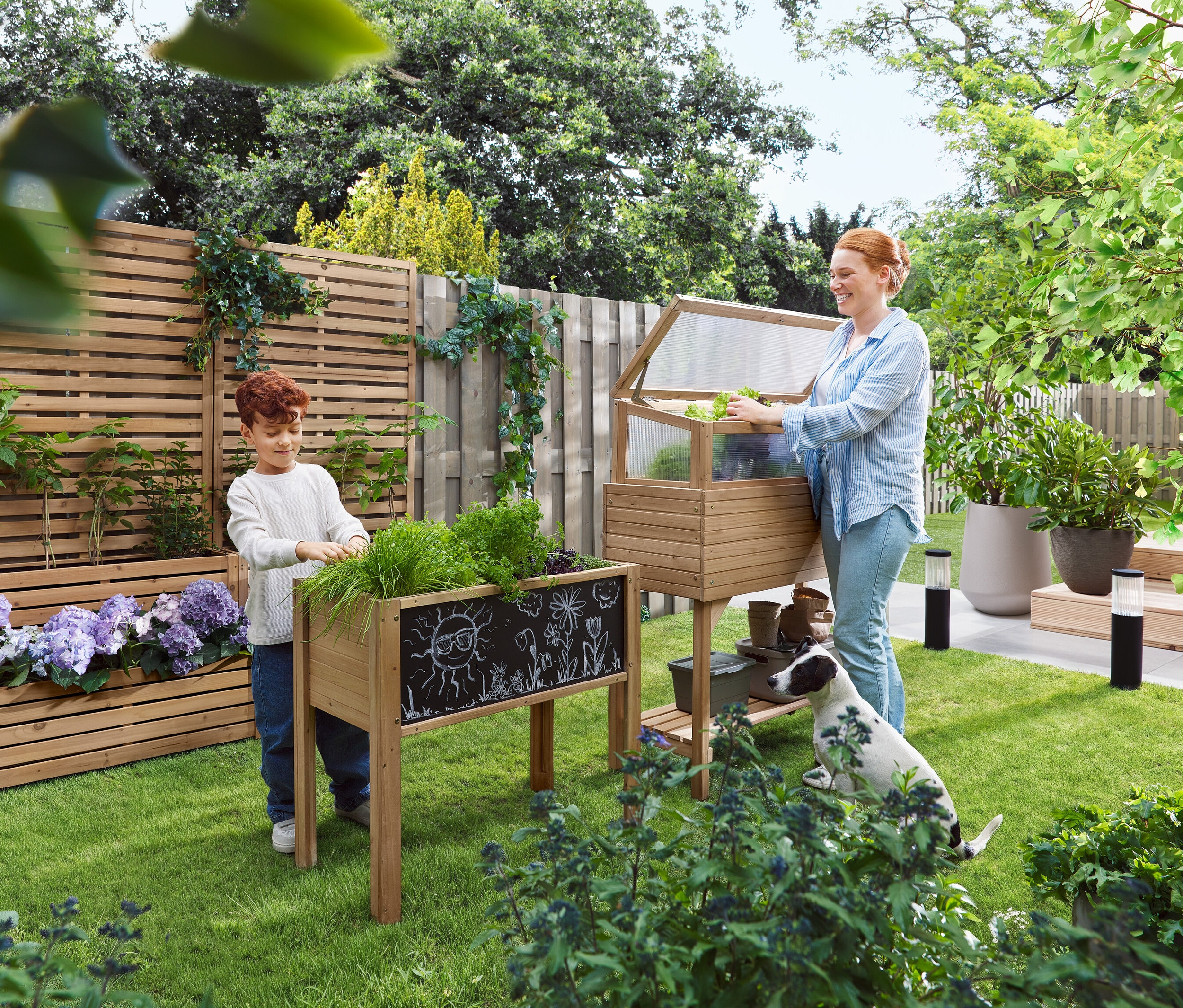 Žena otvára parenisko, zatiaľ čo dieťa stojí vedľa vyvýšeného záhonu. Pes sedí v tráve.