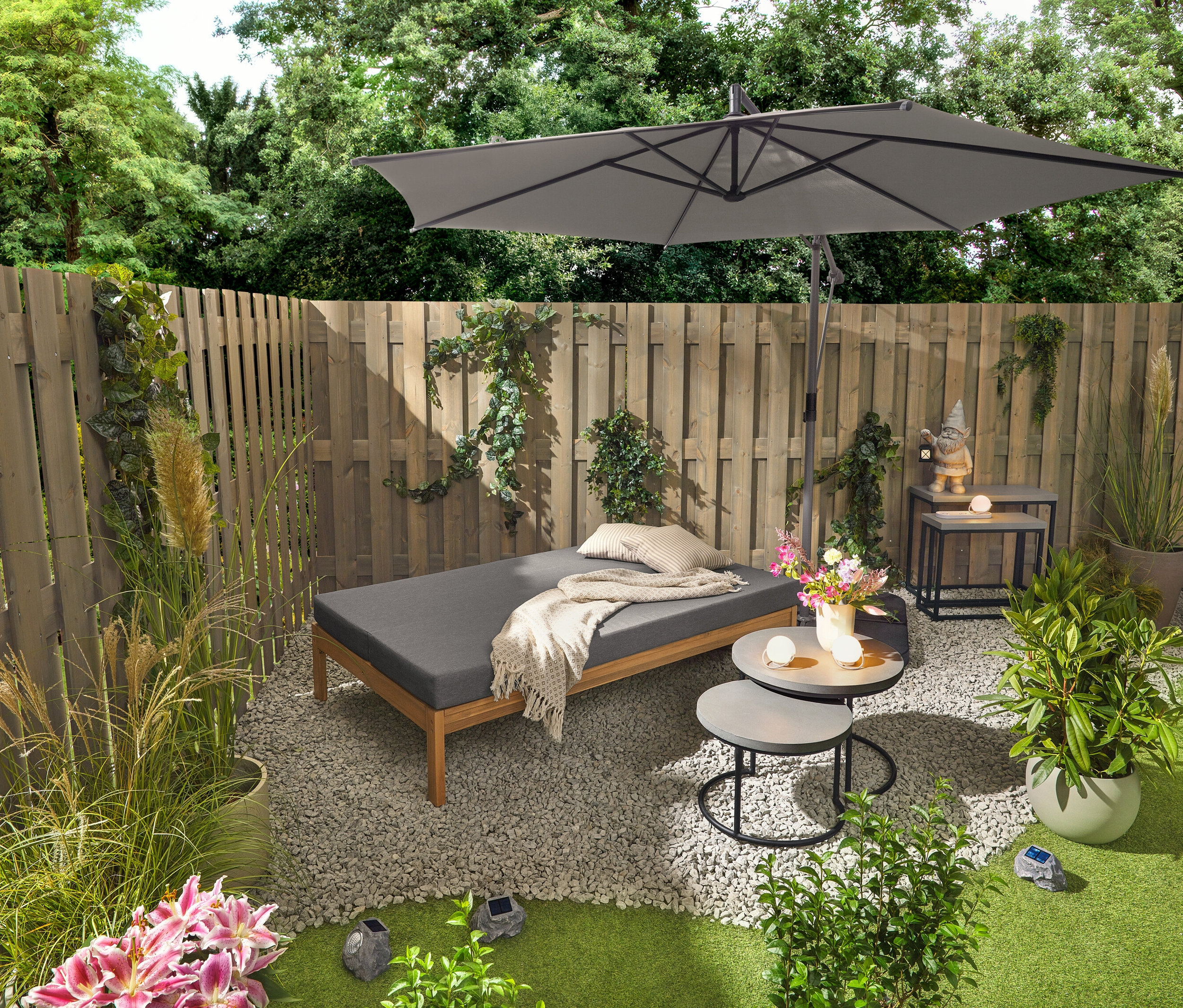 Záhradná lavica 2 v 1, ktorá slúži ako dvojité ležadlo, stojí vedľa dvoch záhradných stolíkov s polycementovými doskami (priemer cca 50 cm a 60 cm). Záhradný trpaslík s LED lampášom stojí na jednom z 2 záhradných konzolových stolíkov s polycementovými doskami. Viditeľné sú aj tri akumulátorové stolové lampy a tri prémiové solárne LED kamene.