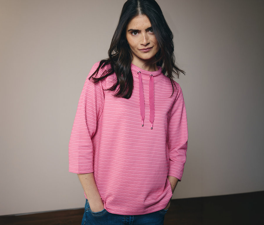 Žena v džínsoch a ružovej mikine CECIL s kapucňou a šnúrkou. Má ruky vo vreckách nohavíc.