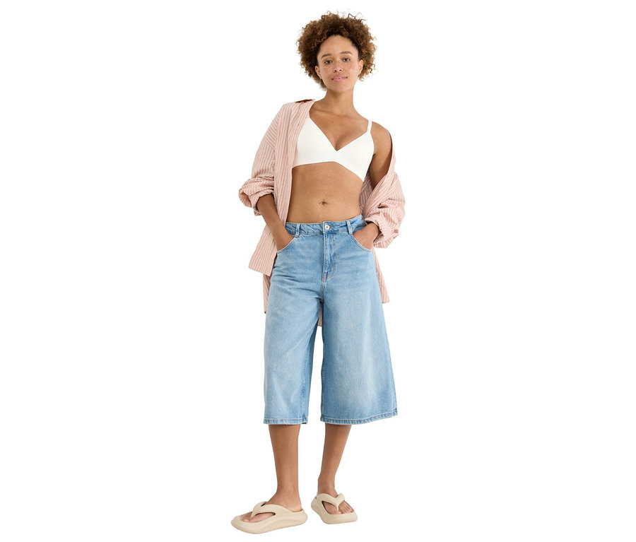 Žena stojí s rukami vo vreckách, má na sebe bielu podprsenku, ružovú pruhovanú blúzku, svetlomodré culottes a béžové žabky.