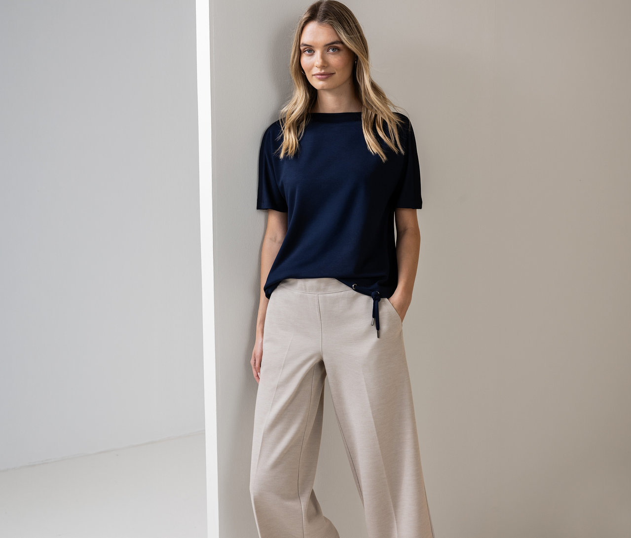 Žena pózuje v námornícky modrom tričku s krátkym rukávom Street One a béžových nohaviciach pri stene.