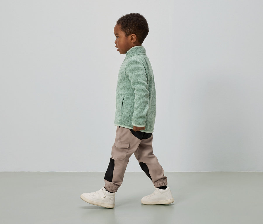 Chlapec s tmavou pokožkou kráča, má na sebe zelenú flísovú bundu, cargo nohavice s čiernymi vložkami a krémové tenisky.