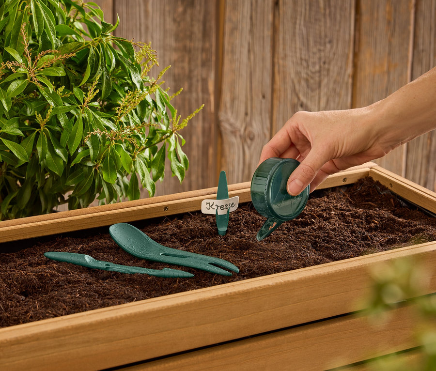 Ruka zalieva vodu z malej zelenej kanvy do drevenej debničky so zemou, v ktorej sú zelené záhradné nástroje a štítok s nápisom "Kresse".