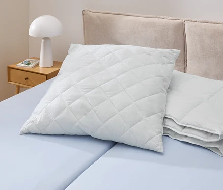 Na svetlomodrej posteli leží biely, prešívaný vankúš Cell Solution® (90 x 70 cm). Vedľa neho sú ďalšie vankúše Cell Solution® (80 x 80 cm, 100 x 65 cm).