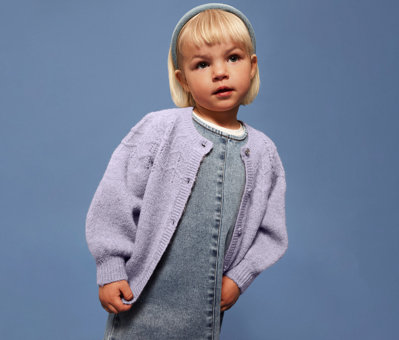 Dievča s blond vlasmi má na sebe svetlofialový detský pletený sveter NAME IT na džínsových šatách na modrom pozadí.
