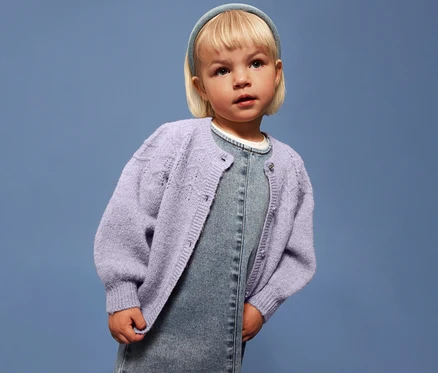 Dievča s blond vlasmi má na sebe svetlofialový detský pletený sveter NAME IT na džínsových šatách na modrom pozadí.