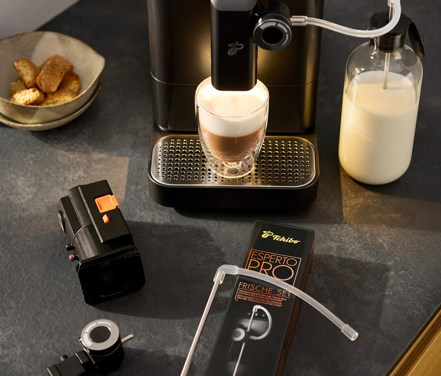 Kávovar »Esperto 2 Milk« Granite Black pripravuje latte macchiato. Vedľa stojí otočný regulátor mliečnej peny, miska s pečivom a balenie súpravy príslušenstva.
