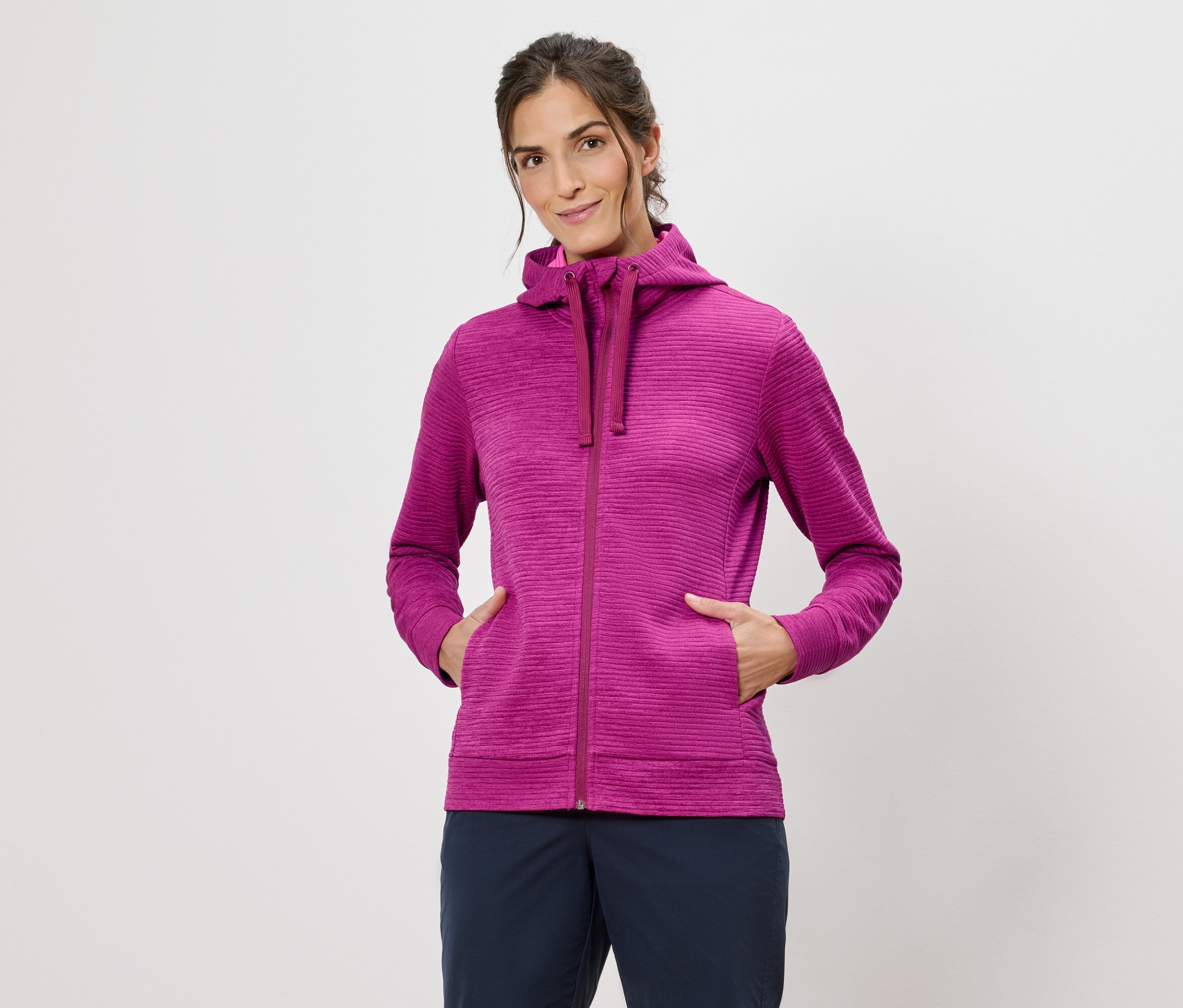 Žena v ružovej mikine s kapucňou s rukami vo vreckách na bielom pozadí.