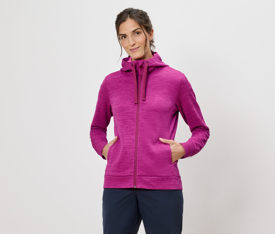 Žena v ružovej mikine s kapucňou s rukami vo vreckách na bielom pozadí.