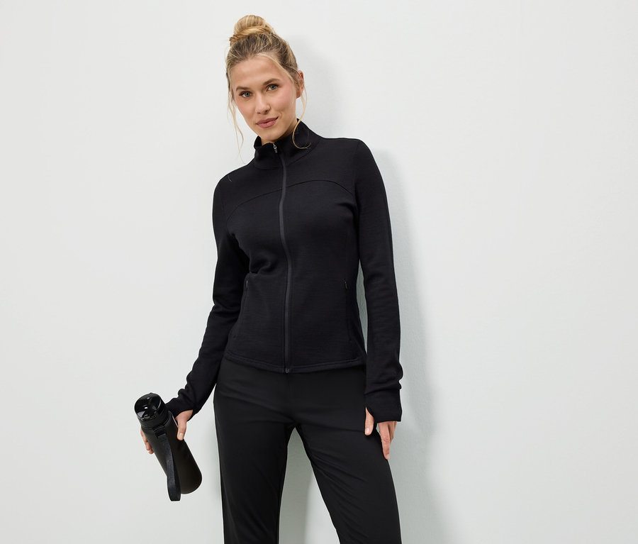 Žena v čiernej bunde a nohaviciach drží čiernu fľašu s vodou na bielom pozadí.