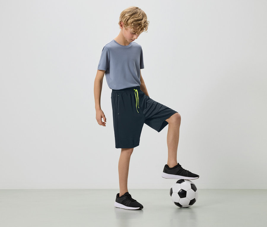 Blond chlapec v sivom tričku a tmavých šortkách s nohou na futbalovej lopte.