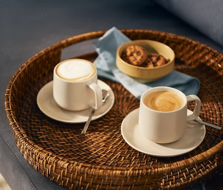 Dve svetlé šálky kávy, jedna s mliečnou penou a druhá s latte art vírom, stoja na zhodných podšálkach s lyžičkami na okrúhlom prútenom podnose. Na podnose je aj malá miska s keksíkmi a svetlomodrá servítka.