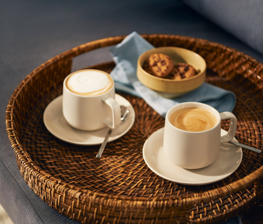 Dve svetlé šálky kávy, jedna s mliečnou penou a druhá s latte art vírom, stoja na zhodných podšálkach s lyžičkami na okrúhlom prútenom podnose. Na podnose je aj malá miska s keksíkmi a svetlomodrá servítka.