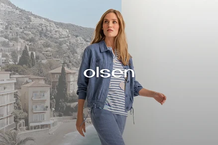 Žena v modrej bunde, pruhovanom tope a modrých nohaviciach, kráčajúca pred rozdeleným pozadím pobrežného mesta a sivej steny, s prekrytým textom 'olsen'.