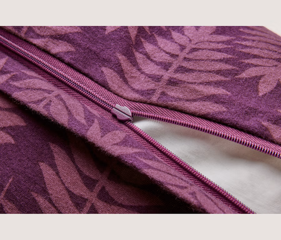 Detail fialovej látky s tmavším listovým vzorom, ukazujúci čiastočne rozopnutý zips rovnakej fialovej farby, pod ktorým je viditeľný biely materiál.