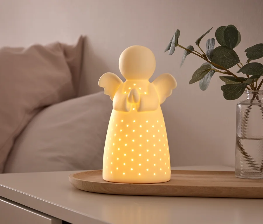Svietiaca biela keramická lampa v tvare anjela s malými perforáciami stojí na drevenom podnose na nočnom stolíku, vedľa vázy so zelenou rastlinou. V pozadí je posteľ so svetloružovými vankúšmi.