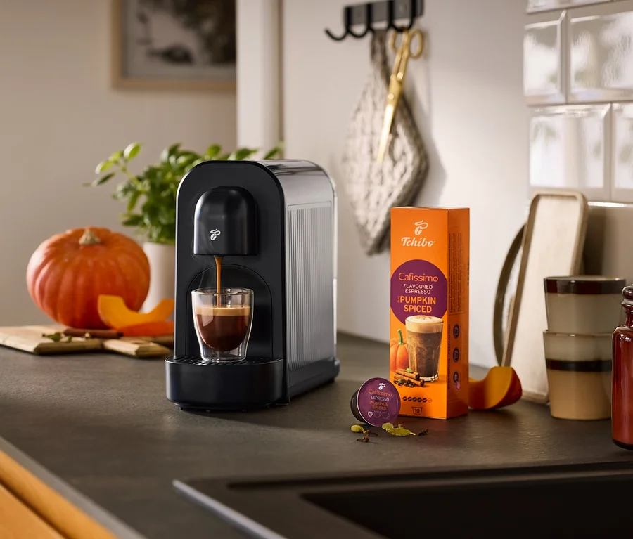 Čierny kávovar Cafissimo now pripravuje espresso do pohára, vedľa neho balenie kávy Cafissimo Pumpkin Spiced a kapsula, obklopené tekvicou a korením na kuchynskej linke.