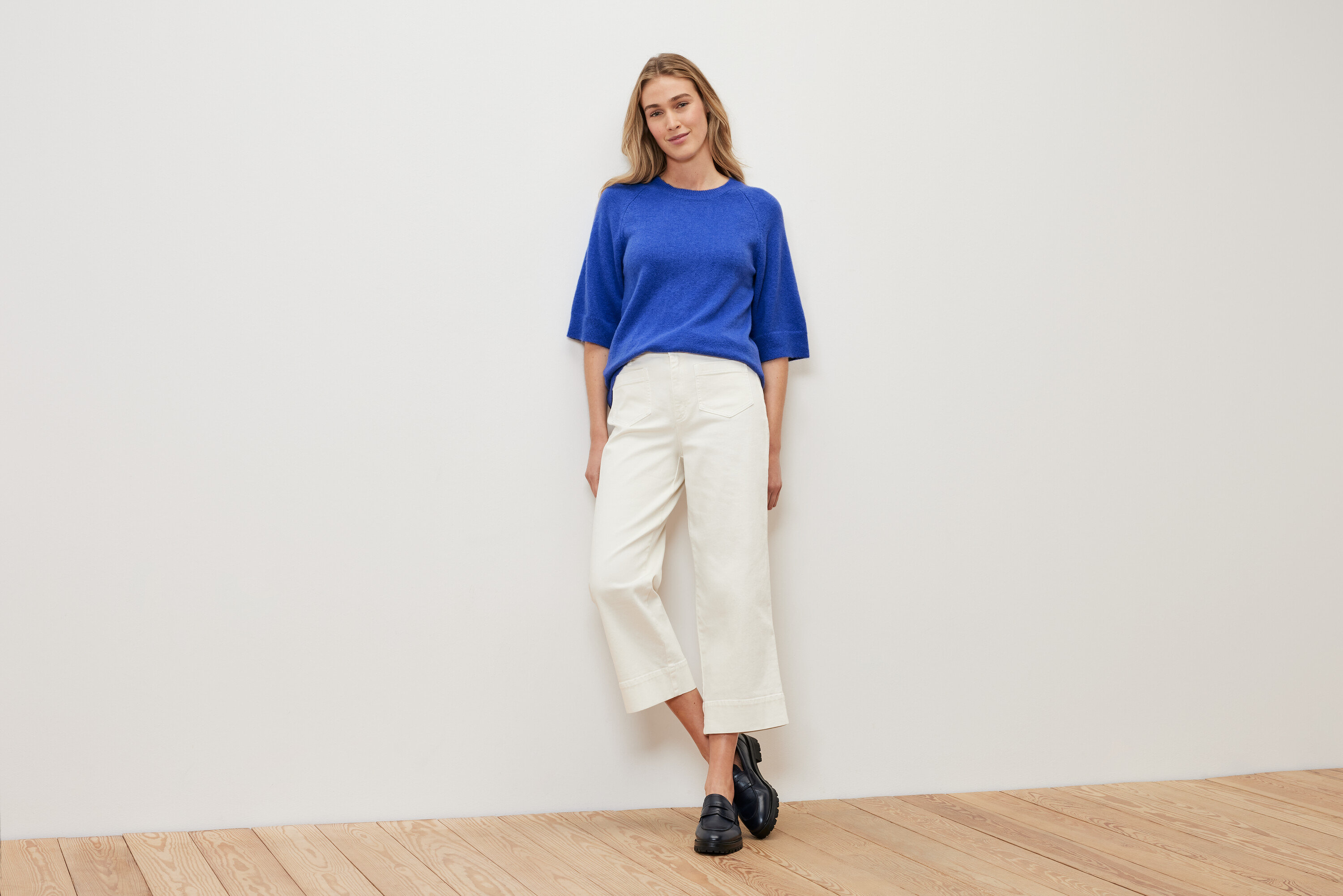 Žena stojaca pred bielou stenou má na sebe pletený pulóver kobaltovomodrej farby, biele nohavice a čierne topánky.