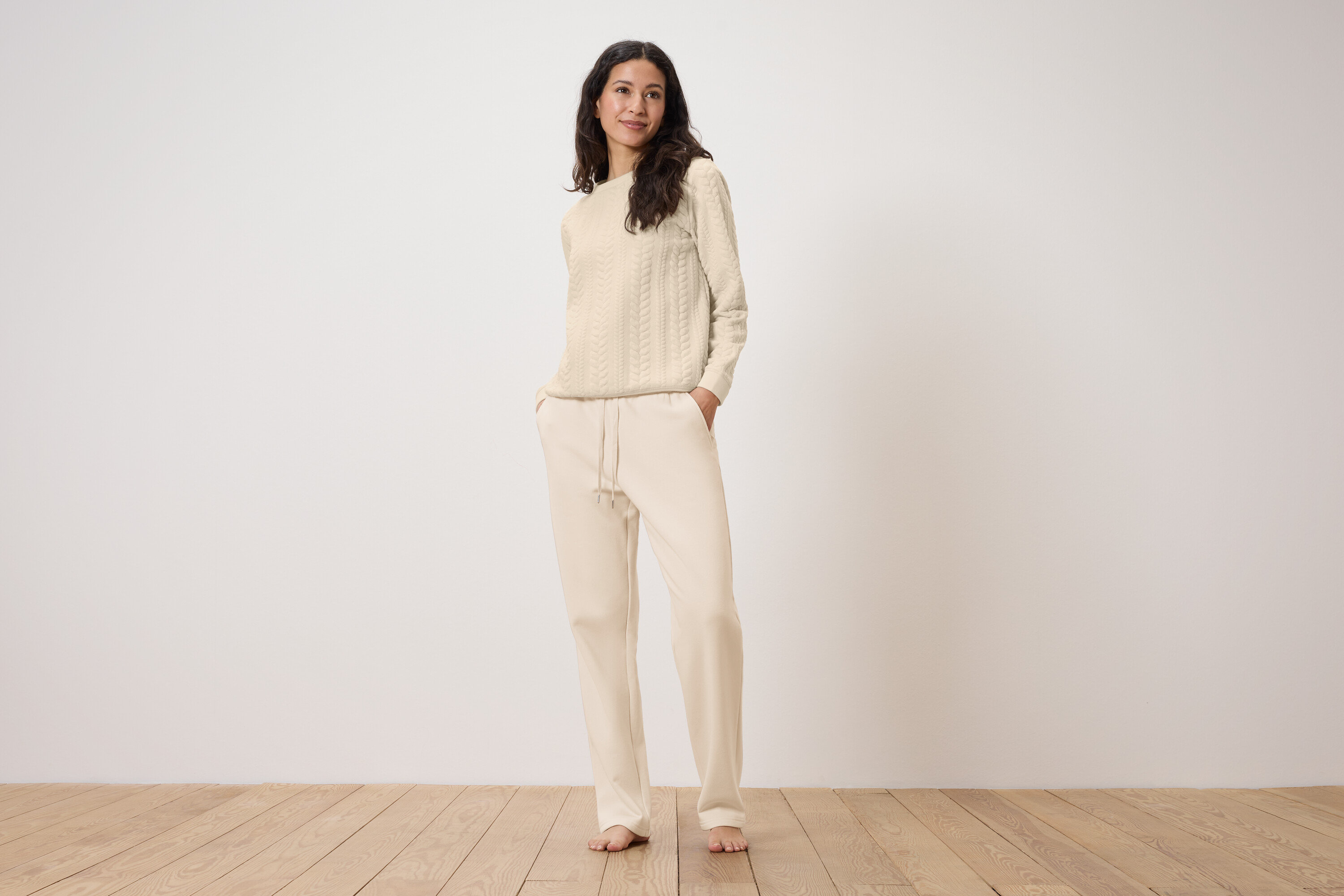 Žena stojí a má ruky vo vreckách krémových teplákov s krémovým svetrom s vrkočovým vzorom.