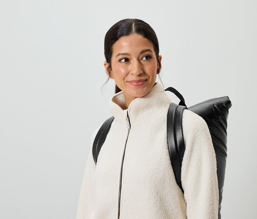 Usmievavá sa žena s hnedými vlasmi a hnedými očami má na sebe krémovú bundu a čierny batoh.