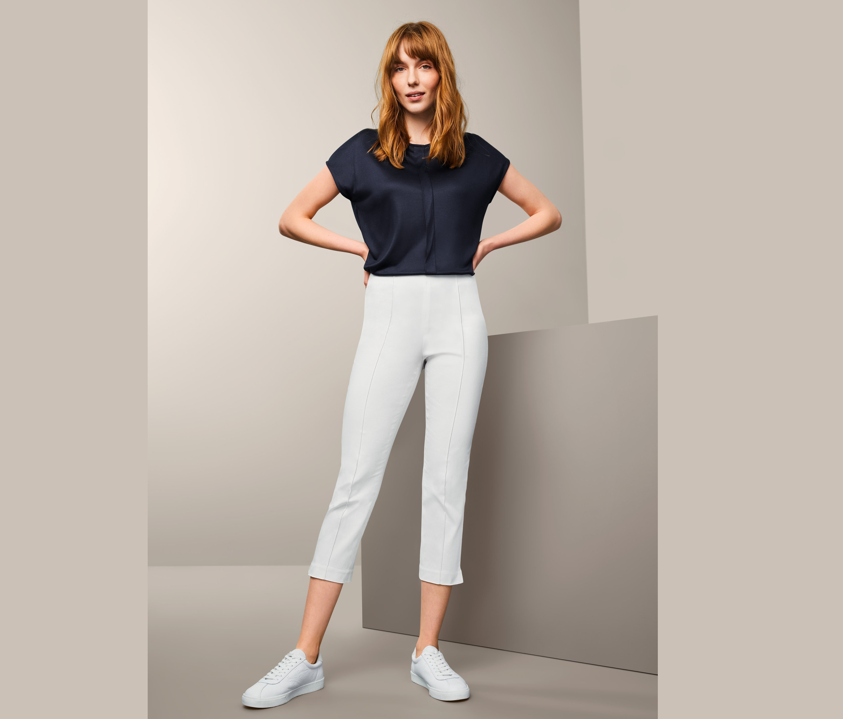 Žena pózuje v bielych sedemosminových elastických nohaviciach a tmavomodrej blúzke.