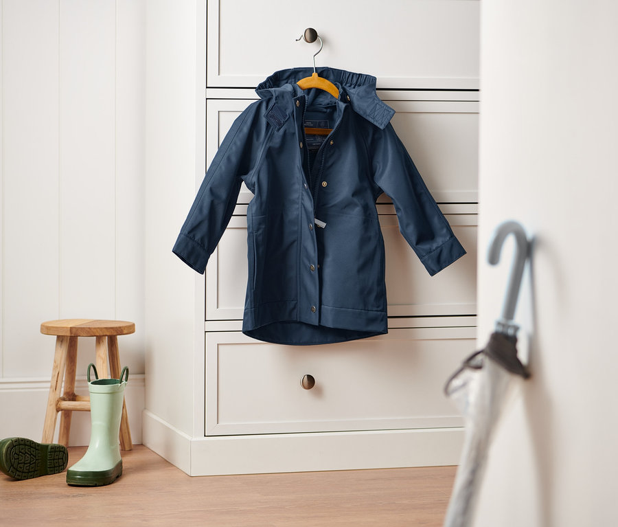 Tmavomodrá bunda do dažďa visí na bielej komode. Na zemi stojí drevená stolička a pár zelených gumákov.