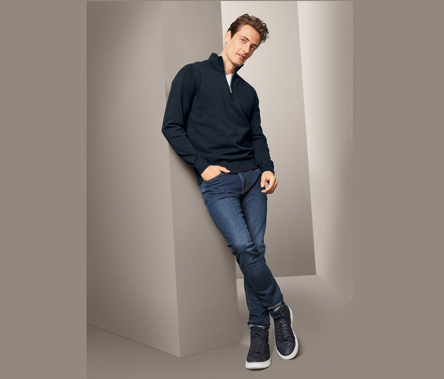 Muž opretý o stenu má na sebe tmavomodrý pulóver z hrubej pleteniny, džínsy a tenisky.