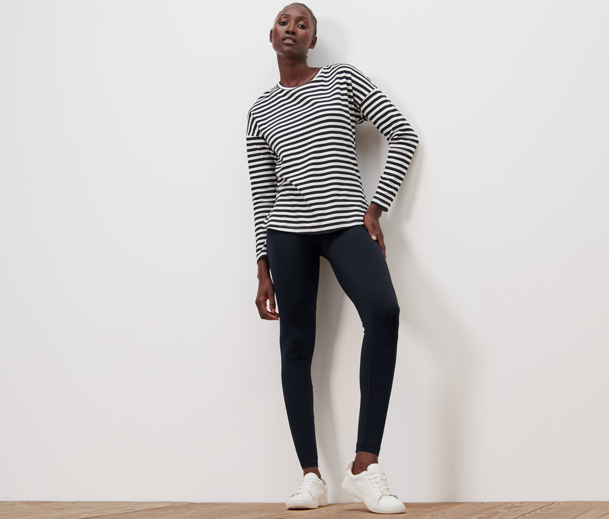 Žena pózuje na drevenej podlahe v čiernych legínach a pruhovanom tričku.