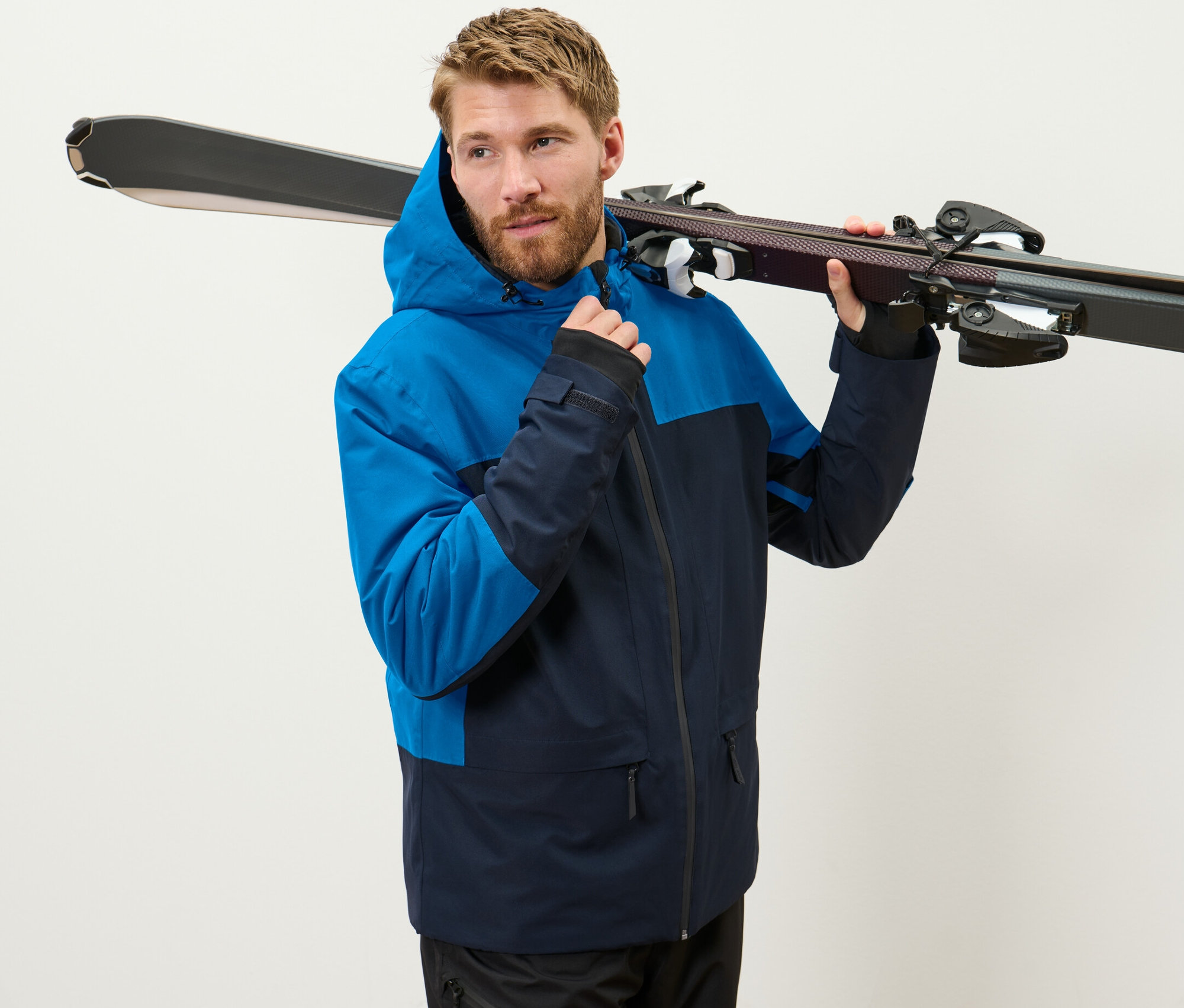 Muž v modrej lyžiarskej bunde drží lyže na ramene.