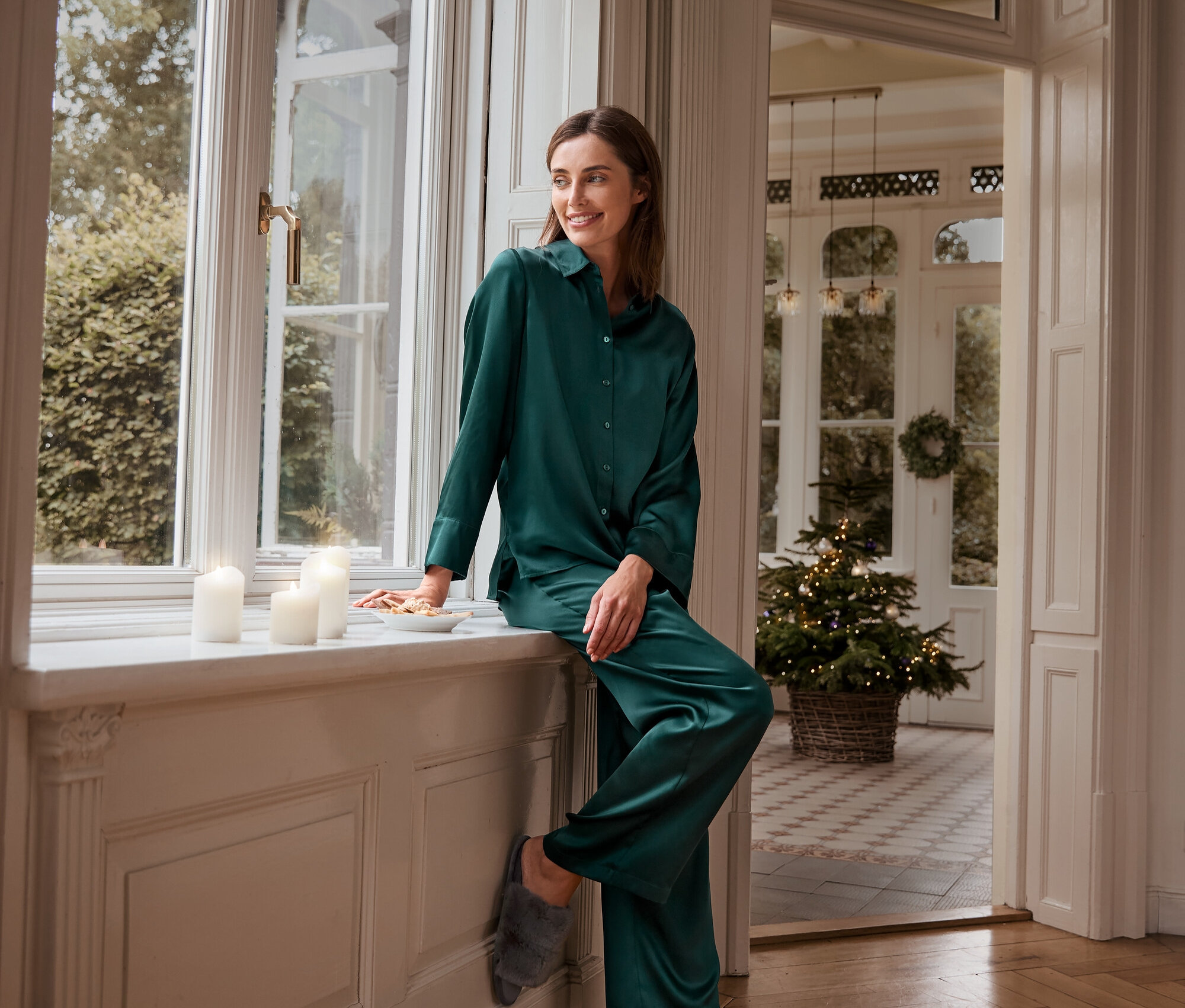 Žena sedí na okennom parapete v zelenom hodvábnom pyžame a plyšových domácich šľapkách.