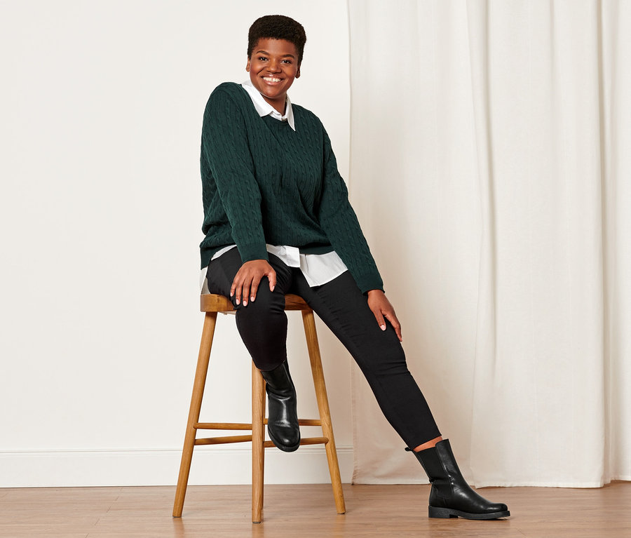 Žena sedí na stoličke a má na sebe tmavozelený pletený pulóver s vrkočovým vzorom, čierne elastické nohavice a kožené topánky.