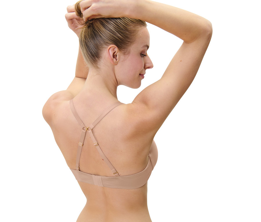 Žena si upravuje vlasy do drdola, na sebe má béžovú podprsenku pod tričko.