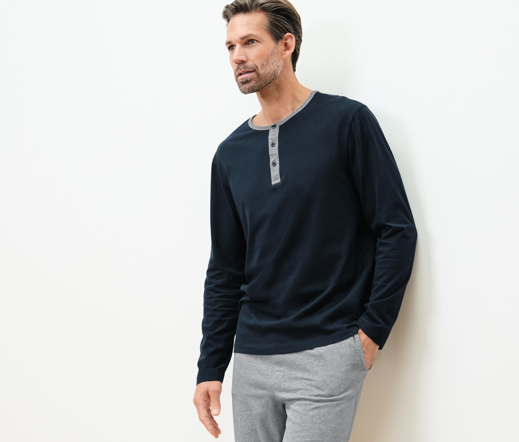 Muž opretý o stenu má na sebe pyžamové tričká s dlhým rukávom (2 ks) a sivé nohavice.