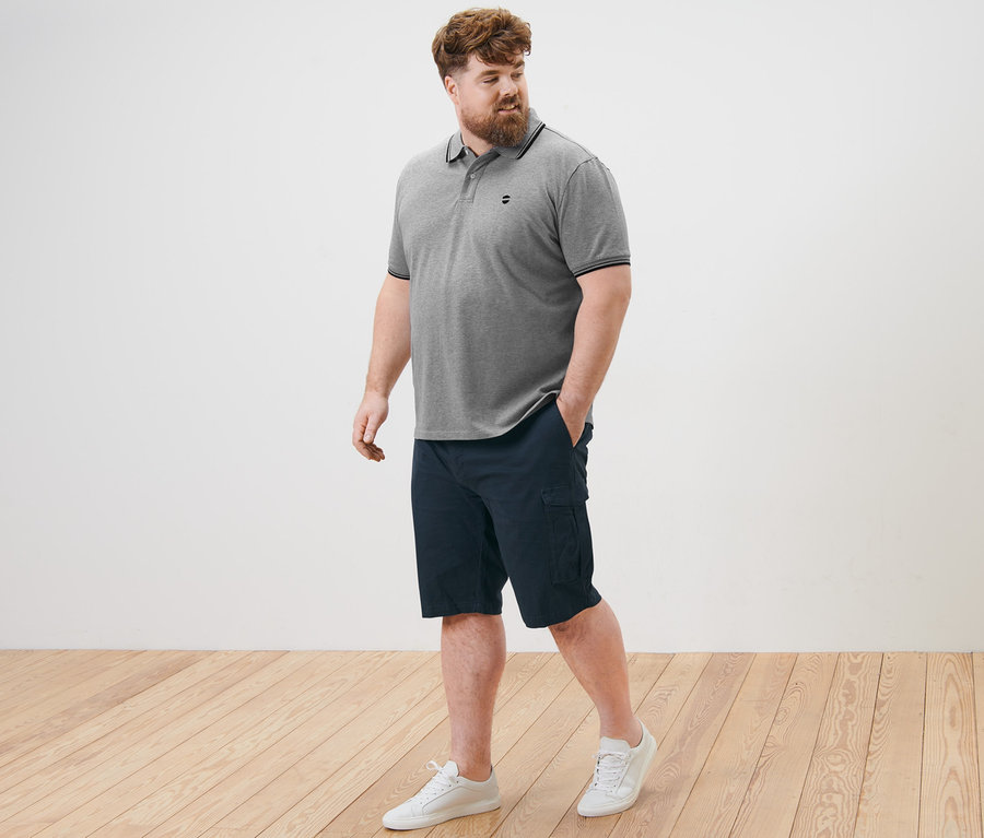 Muž kráča po drevenej podlahe a má na sebe sivú piké polokošeľu a námornícke modré cargo šortky.