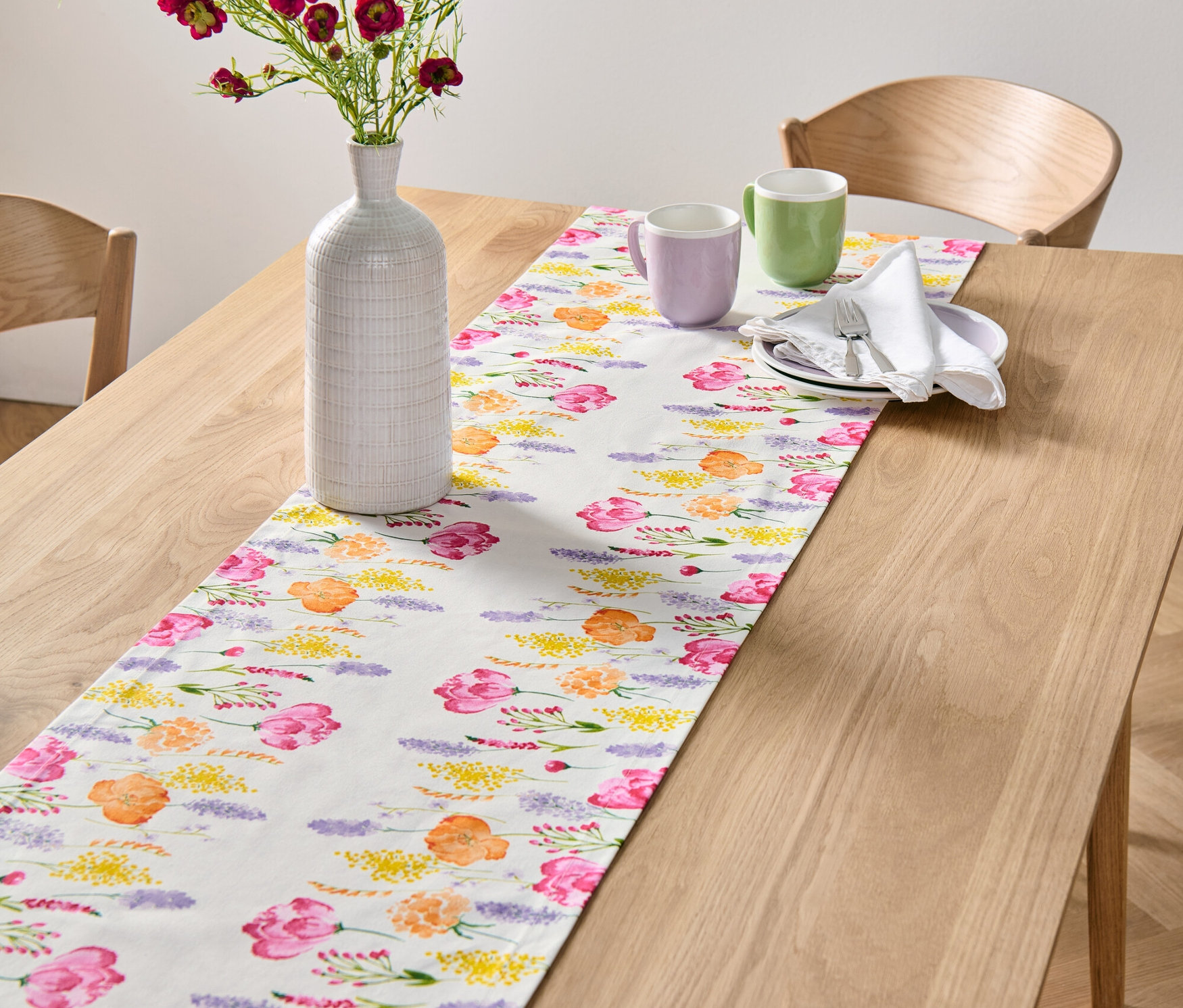 Na drevenom stole je biely behúň s kvetinovou potlačou. Na behúni je váza s kvetmi a dva hrnčeky.