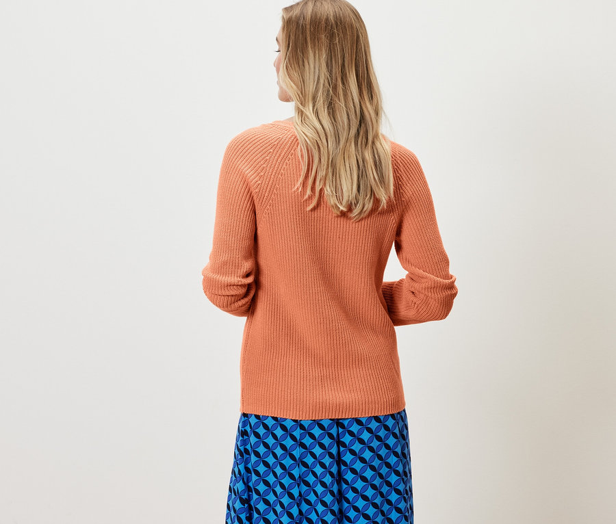 Žena stojí chrbtom k fotoaparátu a má na sebe pletený pulóver broskyňovej farby a modrú sukňu.