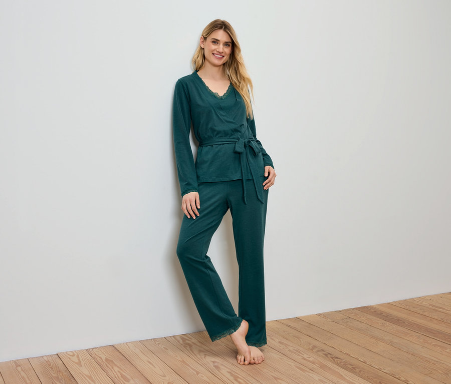 Žena v zelenom pyžame stojí opretá o stenu.