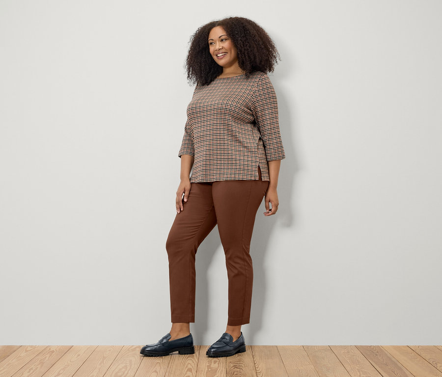 Žena pózuje v hnedej mikine s 3/4 rukávom, hnedých nohaviciach a čiernych topánkach.